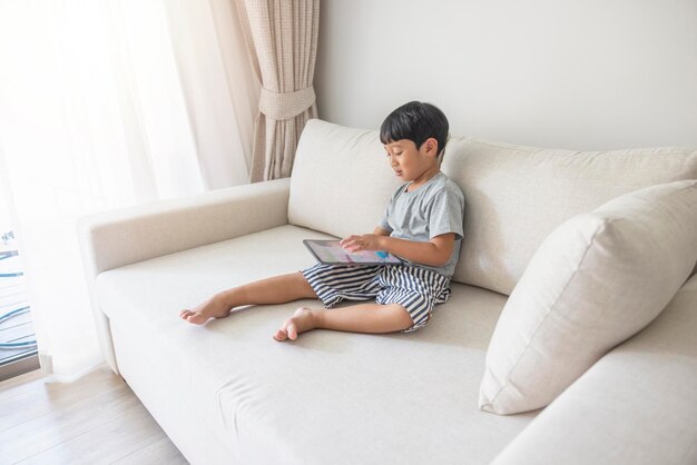 Adorable happy Asian boy wearing a gray shirt and bluewhite striped shorts is having fun playing with his tablet on a cream sofa looking at the mobile screen