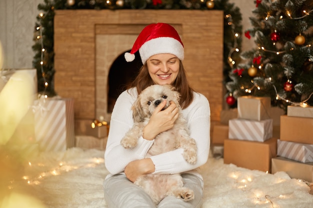 Adorabile ragazza felice in cappello rosso che abbraccia con simpatico cane pechinese da albero di natale con luci, camino e scatole regalo nella stanza festiva, la femmina si siede sul pavimento e si diverte con il cucciolo.