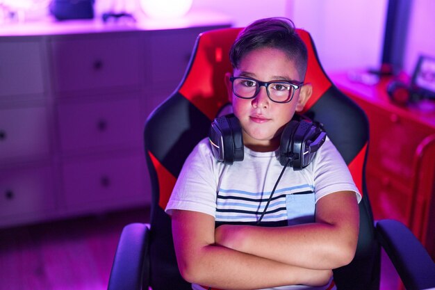 Adorable hispanic boy streamer sitting with arms crossed gesture at gaming room