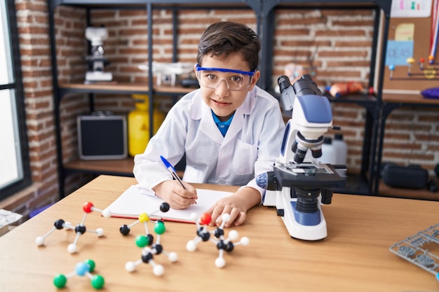 Adorable hispanic boy student using microscope writing on notebook at laboratory classroom