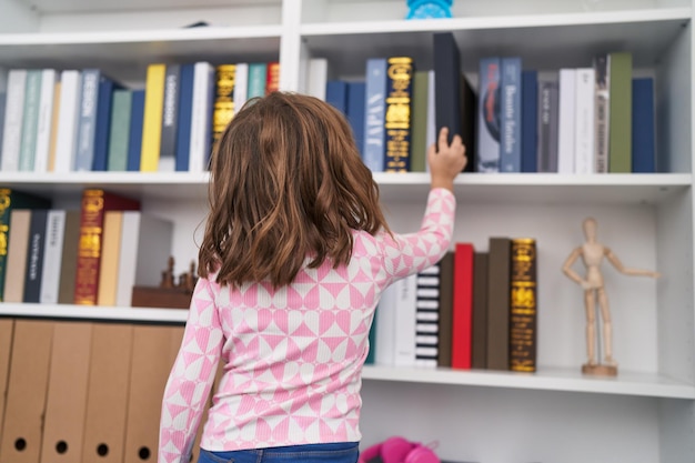 Photo adorable hispanic girl student holding book of shelving at library school