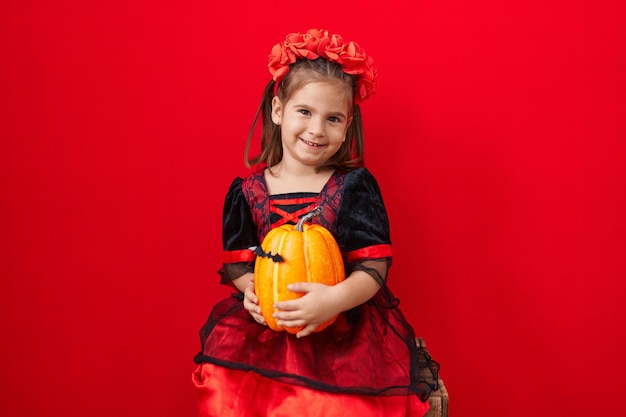 Adorable hispanic girl wearing halloween costume holding pumpkin over isolated red background