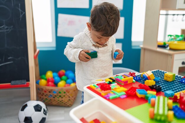 Adorable hispanic toddler playing with construction blocks standing at kindergarten