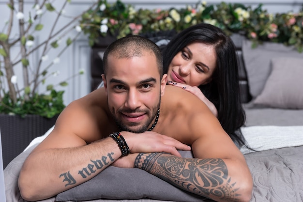 Adorable international couple of man with bare chest and with tattooed hands, brunette woman lying on him on the gray cozy bed in the bedroom