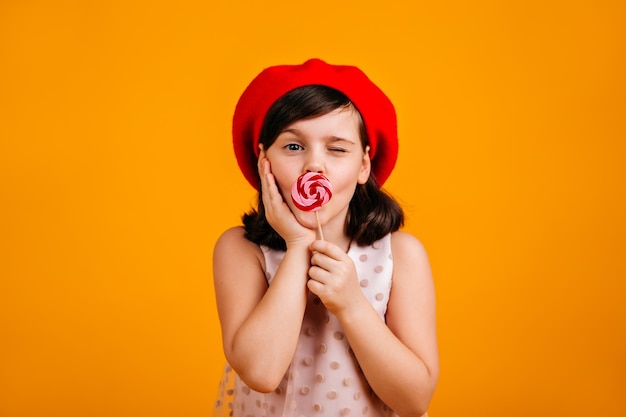 喜びでキャンディーを食べる愛らしい子供黄色の背景に分離されたロリポップとブルネットの少女のスタジオショット