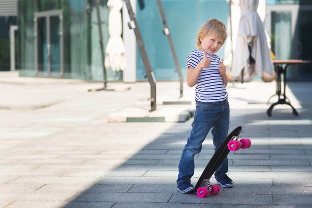 Adorable kid outdoors. Cute pretty child smiling at camera. Casual boy on summer time skating on a skateboard. thumb up