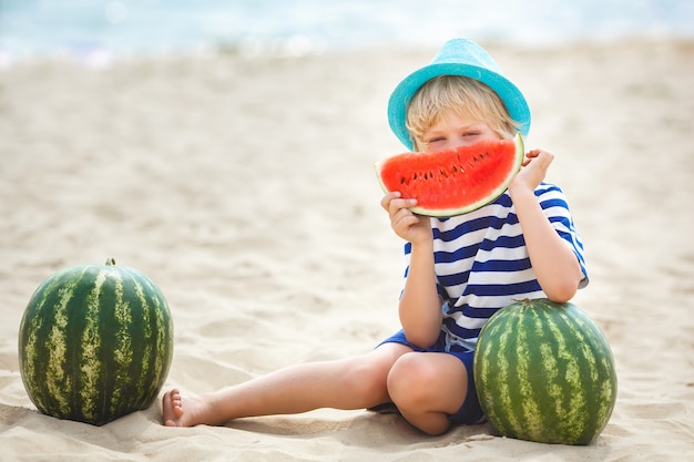 Прелестный ребенок на берегу моря, едят сочный арбуз. Веселый ребенок на летнее время на пляже. Милый маленький мальчик на улице