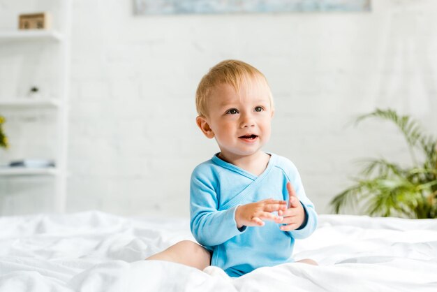 Adorable kid sitting on bed with white bedding and gesturing at home