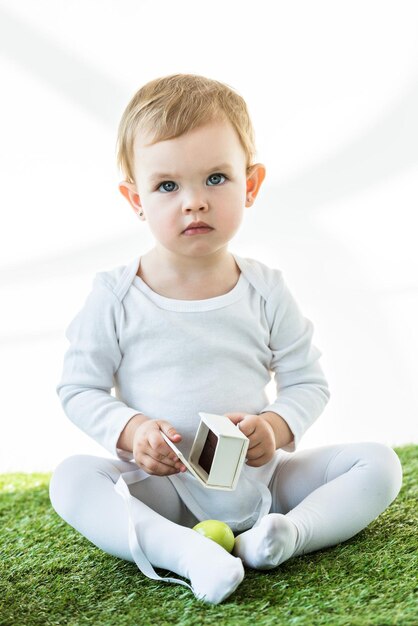 Adorable kid sitting on green grass with white cardboard box and yellow chicken egg isolated on