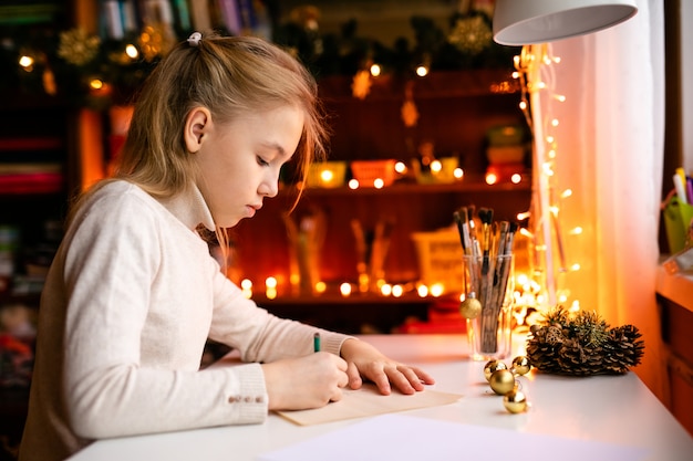 Adorable litle girl is writting a letter to santa klaus sitting on the table