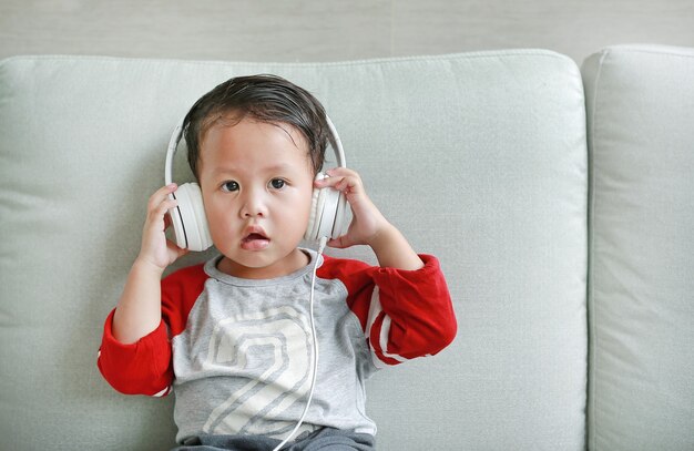 Adorable little Asian baby boy in headphones is using a smartphone lying on the sofa at home. Child listening to music on earphones.
