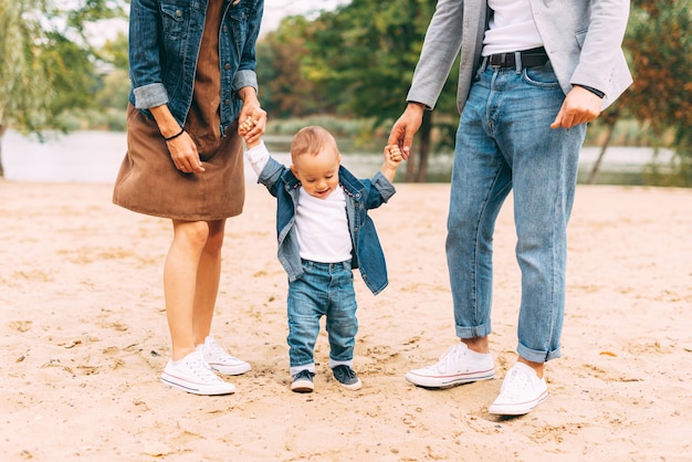 adorable little baby is learning how to walk near his parents on sand near a lake