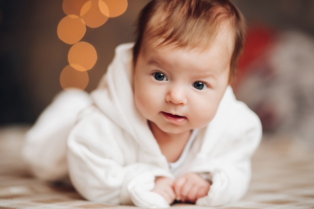 Photo adorable little baby with fair hair in white body with hood lying on the bed under snowfall