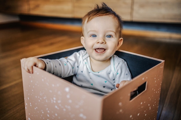 Adorable little blond boy with blue eyes sitting in box, laughing and playing