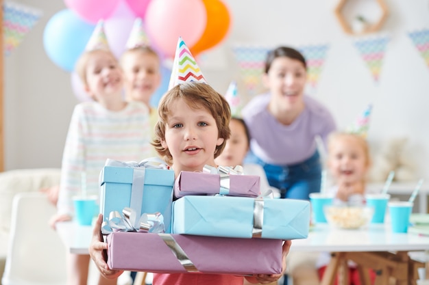 Adorable little boy in birthday cap holding pile of birthday presents at home party with friends