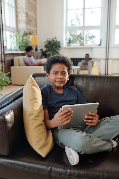 캐주얼한 옷을 입은 사랑스러운 소년은 베개 옆에 검은 가죽 소파에 앉아 가제트를 사용하여 부모에 대해 태블릿으로 온라인 비디오를 보고 있습니다.