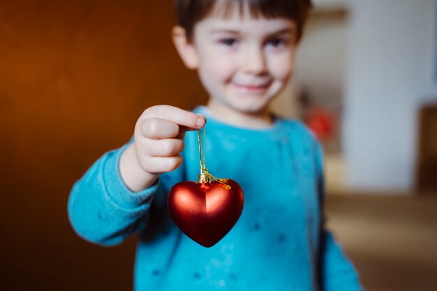 Adorable little boy holding with cheerful smile a red shaped heart on her living room