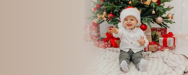 Adorable little boy in a New Year's hat sits on a blanket in front of the Christmas tree