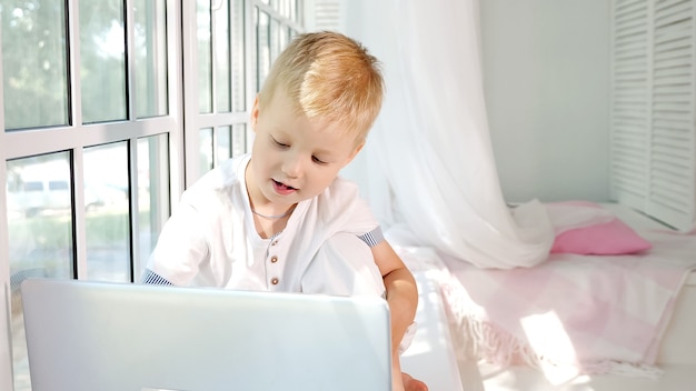 Adorable little boy sitting on sill of big white window in laptop