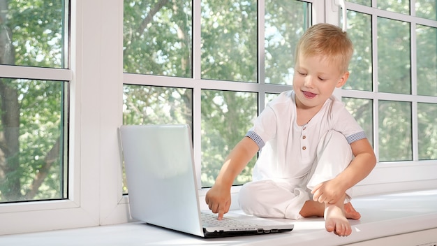 Adorable little boy sitting on sill of big white window in laptop