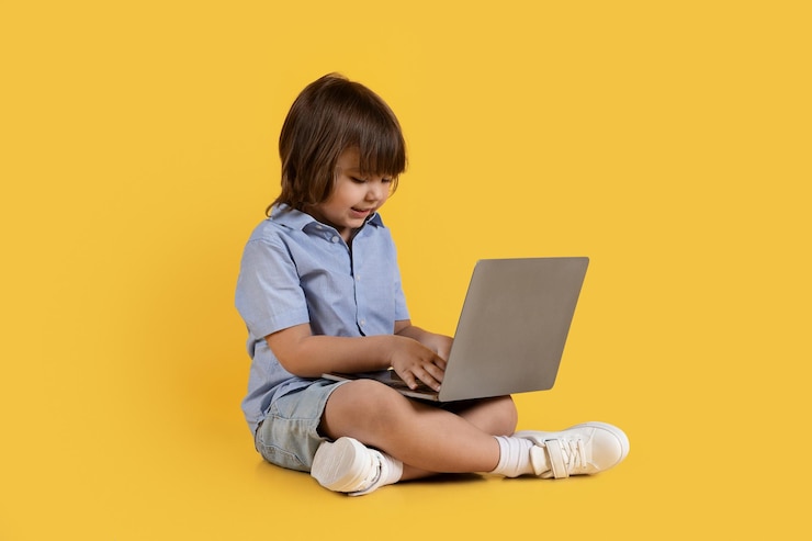 Photo adorable little boy typing on laptop computer playing developing game sitting on floor orange studio background
