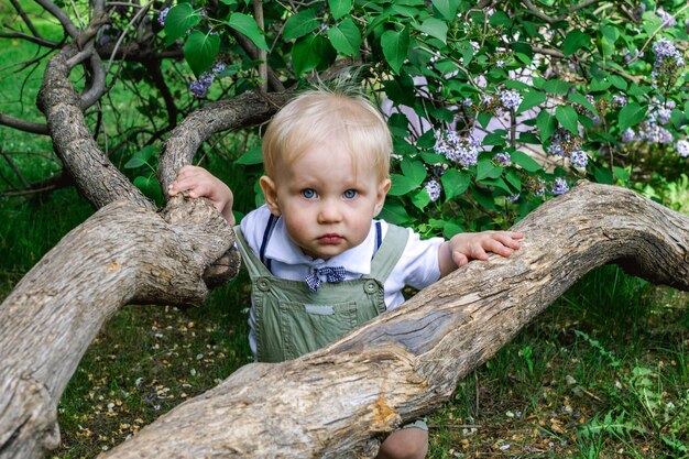 Photo adorable little boy with blue eyes near to tree in blossom spring garden with purple lilac flowers