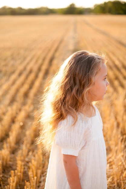 Adorable little curly girl 4 years old in a white dress in the sun at sunset in a mowed field of wheat Happy child outside Walk Warm summer Emotions