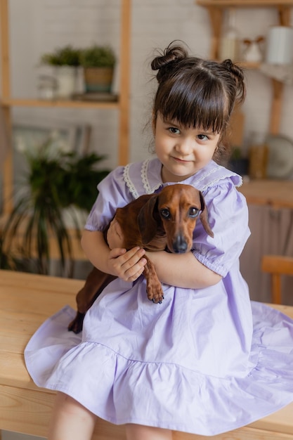 Adorable little girl in a beautiful dress plays with a dachshund dog in the kitchen and feeds her