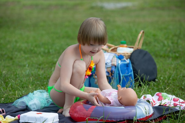 Adorable little girl in a bright swimsuit plays with her favorite doll during a picnic in the park