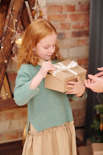 Adorable little girl in casualwear opening giftbox with Christmas present from her mother while standing by brick wall in home environment