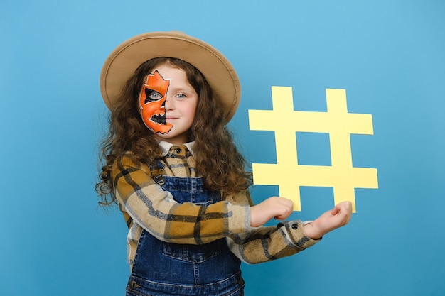 Foto adorabile bambina con maschera di halloween che tiene il simbolo dell'hashtag giallo e felice guardando la telecamera, mostrando il segno di hash, isolato su sfondo blu. post alla moda sui social media e concetto di festa