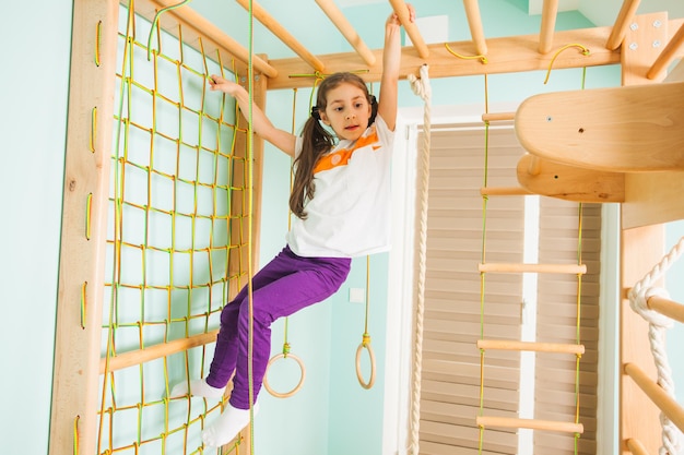 Adorable little girl climbing wooden bars and rope ladder at home sport gym. Small future gymnast girl training at home