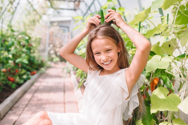 Adorable little girl harvesting cucumbers and tomatoes in greenhouse.