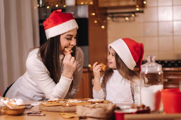 Adorable little girl and her mom in santa hat eating homemade gingerbread and laughing happy little