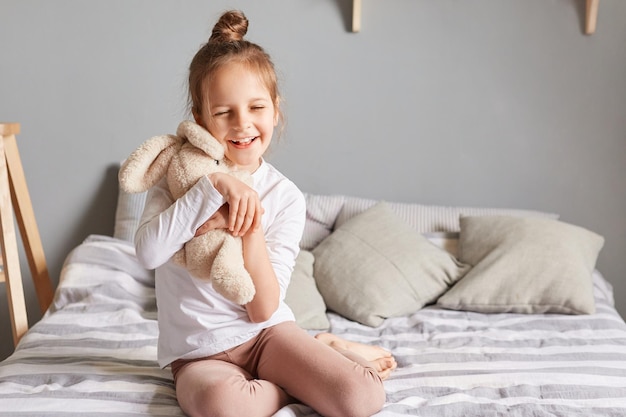 Adorable little girl in home clothing playing with fluffy rabbit kid having fun with soft toy child playing with hare embracing favorite bunny smiling happily