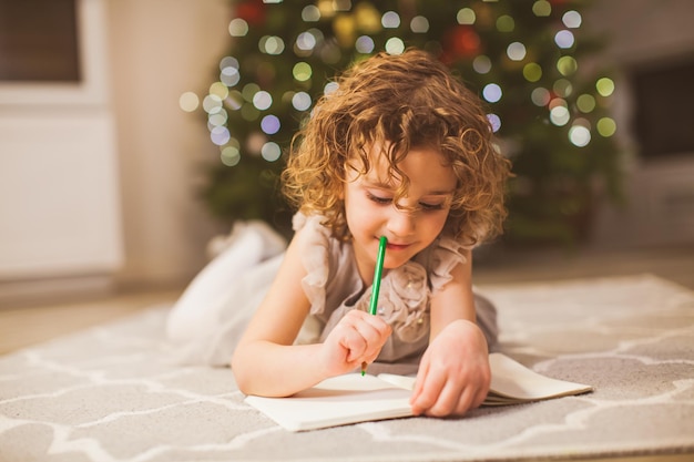 背景にサンタクロースのクリスマスツリーに手紙を書くことを考えている彼女の口に緑の鉛筆を保持している柔らかいカーペットで床に横たわっているお祝いのドレスを着た愛らしい少女