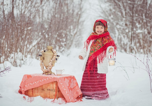 Очаровательная маленькая девочка в старой одежде в зимнем лесу с самоваром и рогаликами