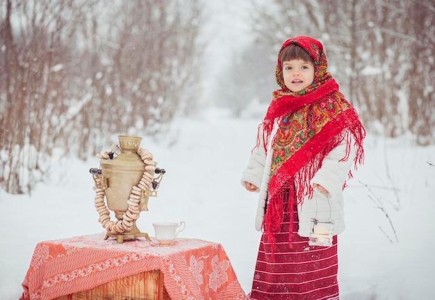 Adorable little girl in old Russian clothes in a winter forest with a samovar and bagels