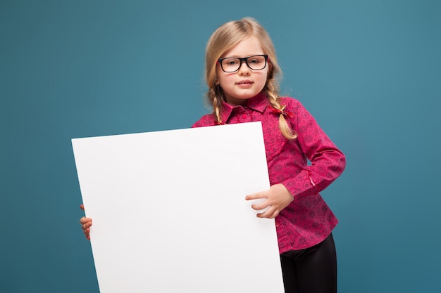 La bambina adorabile in camicia rosa, pantaloni neri e vetri tiene il cartello in bianco vuoto