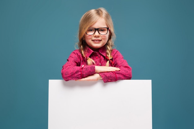 Adorable little girl in pink shirt, black trousers and glasses holds empty blank placard