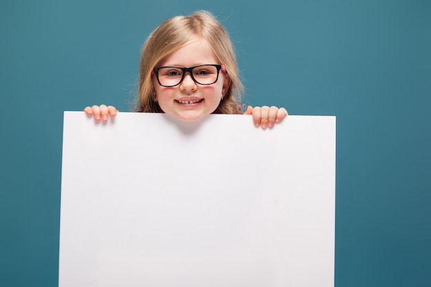 Adorabile bambina in camicia rosa, pantaloni neri e occhiali detiene poster vuoto