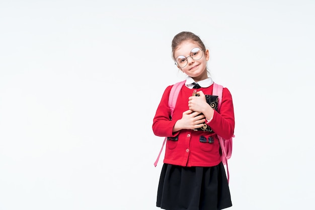 La bambina adorabile in rivestimento rosso della scuola, vestito nero, zaino e vetri arrotondati abbraccia strettamente un libro e sorride mentre gode e posando sullo spazio bianco. isolato