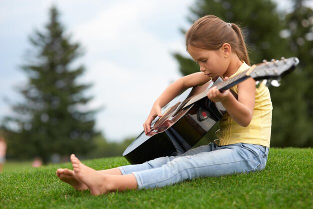 Adorable little girl sitting on a green grass in park and holding guitar spending time with parents outdoors