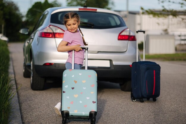 Adorable little girl standing in front of the car with her kid suitcase and waiting for parents