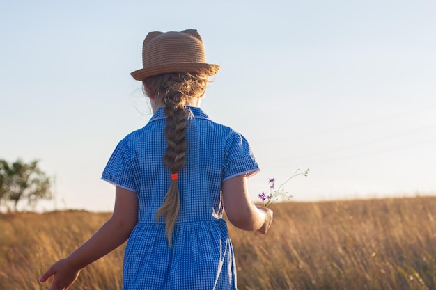 Adorable little girl in a straw hat blue plaid summer dress in grass field countryside Child with long blonde braid