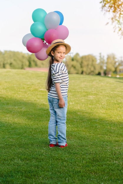 Adorable little girl in straw hat holding colorful balloons and smiling at camera