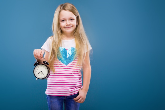 Adorable little girl in white striped shirt with blue heart and jeans hold alarm clock