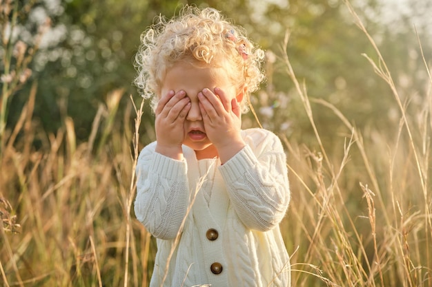 Adorable little girl in white sweater covering eyes with hands while standing on grassy meadow in sunny summer countryside