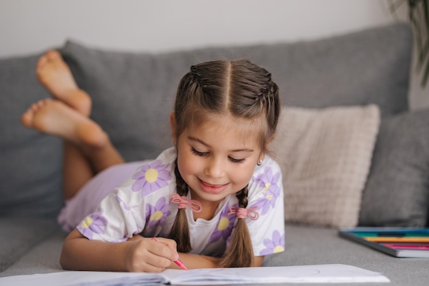 Adorable little girl with beautiful braid hairstyle lying on sofa at home and drawing picture for