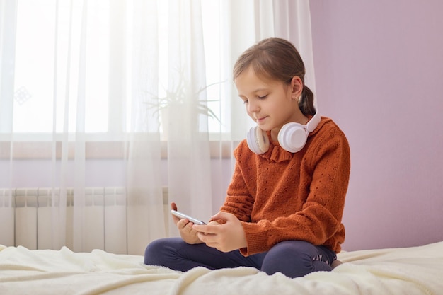 Photo adorable little girl with brown hair sitting on bed using mobile phone wearing headphone over neck using smartphone app communicating with friends browsing internet pages